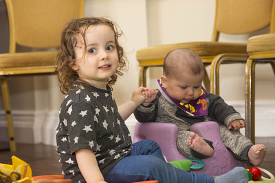 Two babies sitting on the floor, holding hands. One of them is looking and smiling at the camera; the other is looking away.